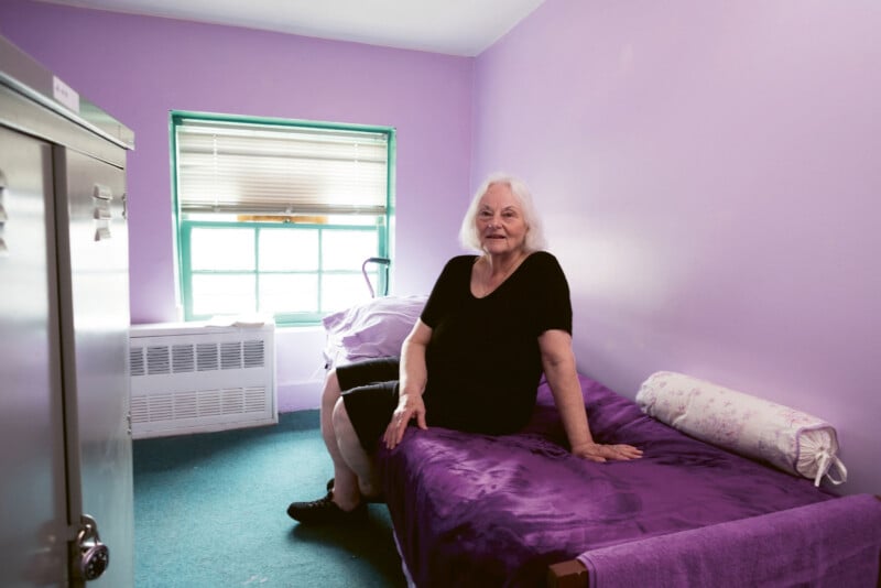 An older woman with white hair sits on a purple-covered bed in a small room with lavender walls and green carpet. A window lets in daylight and there is a pillow and blanket on another bed nearby.