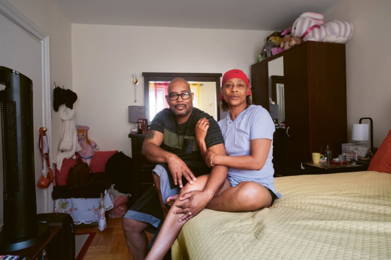 A couple sits close together on the edge of a bed in a cozy bedroom, smiling at the camera. The room has stuffed animals on top of a wardrobe and various personal items on the dresser.