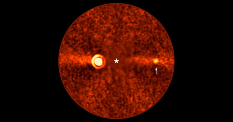 Orange-hued, grainy circular image of a star at the center with a glowing ring to the left and a smaller bright spot to the right, indicated by an arrow. The background is dark with scattered noise.