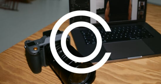 A digital camera rests on a wooden table next to an open laptop displaying photo editing software. A large white copyright symbol overlays the image, partially obscuring the view.