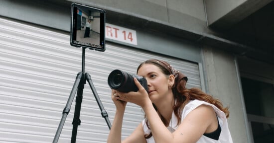A woman holding a camera prepares to take a photo, next to a tripod-mounted tablet displaying a live preview of her shot, in front of a metal garage door.