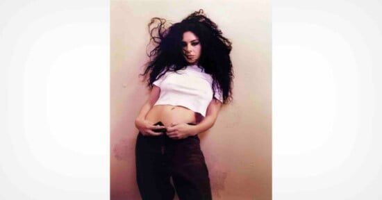 A person with long, curly dark hair in a white crop top and dark pants stands against a light-colored wall, looking at the camera with a serious expression.