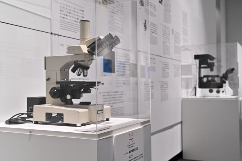 A Nikon microscope is displayed inside a glass case on a pedestal in a museum, with another microscope visible in the background.