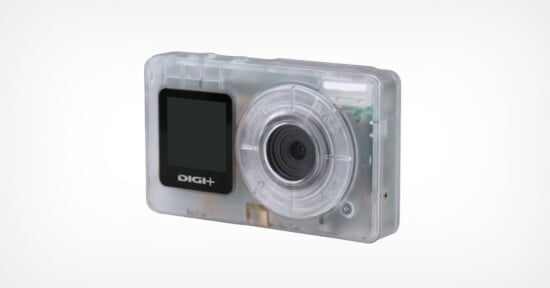 A compact digital camera with a transparent plastic case, revealing internal components. The front features a lens on the right and a small display screen labeled "DIGI+" on the left.
