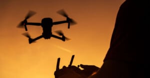 A person operates a drone with a remote controller at sunset, the drone flying against an orange sky.