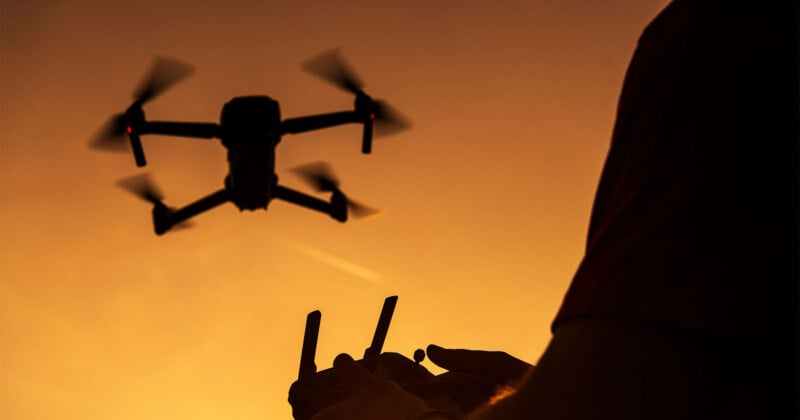 A person operates a drone with a remote controller at sunset, the drone flying against an orange sky.