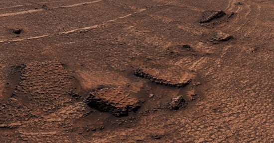 A high-angle view of a reddish, arid Martian landscape featuring polygonal patterned ground and several rocky outcrops.