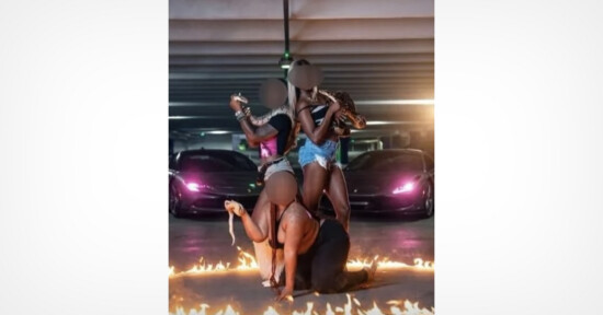 Three women pose in a parking garage surrounded by a ring of fire; two stand back to back, while one kneels in front. Two sports cars with glowing headlights are parked behind them. Faces are blurred.