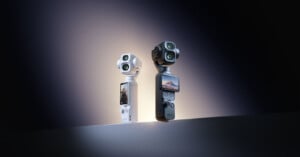 Two DJI Osmo handheld gimbal cameras, one white and one black, stand upright on a flat surface against a dark, gradient background.