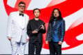 Three men stand on stage in formal attire, one in a white tuxedo, one in a black suit holding an award, and one in a blue patterned jacket. A red-and-white abstract background is behind them.