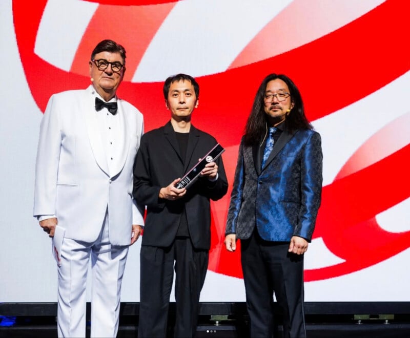Three men stand on stage in formal attire, one in a white tuxedo, one in a black suit holding an award, and one in a blue patterned jacket. A red-and-white abstract background is behind them.
