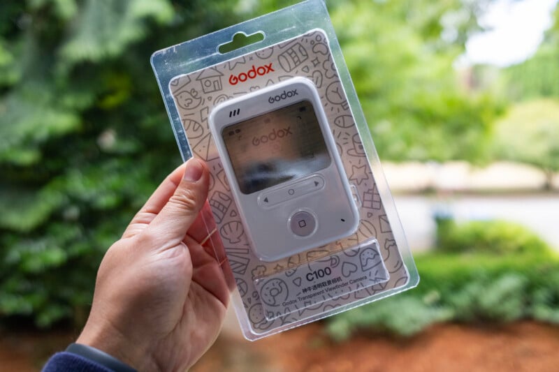 A hand holds a packaged Godox C100 portable device outdoors, with greenery and a path blurred in the background.