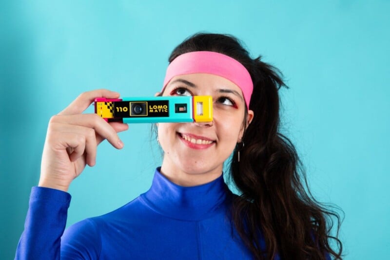 A person with a pink headband and blue long-sleeved shirt holds a colorful "Lomo Matic" 110 camera in front of their eyes.