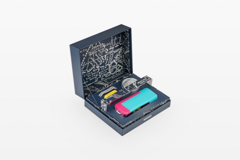 An open blue gift box with a map-patterned interior contains a bright pink and blue rectangular device, a small silver coin, and a yellow coiled cable.