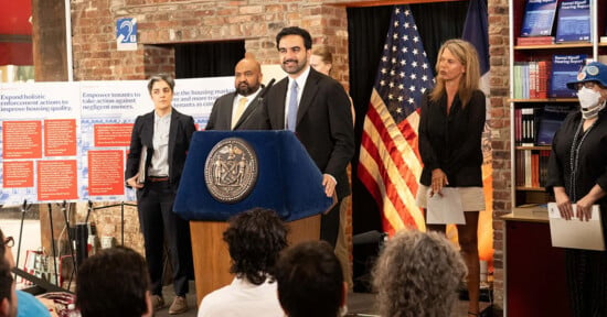 A man speaks at a podium with a city seal, flanked by officials. Behind them are American flags, posters with text, and a brick wall. People in the audience listen attentively.
