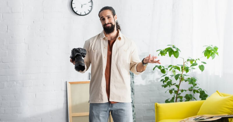 A man with a beard holds a camera in one hand and shrugs with the other, standing in a bright room with a yellow couch, green plant, and a clock on the wall.