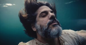 A bearded man with closed eyes and flowing hair floats underwater, wearing a tattered light shirt, appearing serene and calm.