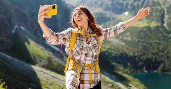A smiling woman with wavy hair and a yellow backpack wearing a plaid shirt takes a selfie with her yellow phone while standing on a mountain trail with arms wide open, overlooking a scenic valley with a lake below.