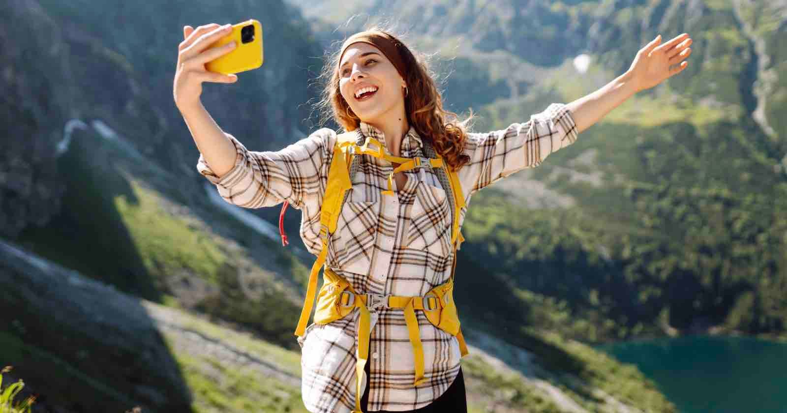 A smiling woman with wavy hair and a yellow backpack wearing a plaid shirt takes a selfie with her yellow phone while standing on a mountain trail with arms wide open, overlooking a scenic valley with a lake below.