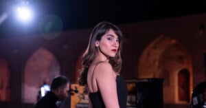 A woman in a black dress stands in focus, looking over her shoulder at the camera. She is outdoors at night, with arched brick architecture and soft lighting in the background.