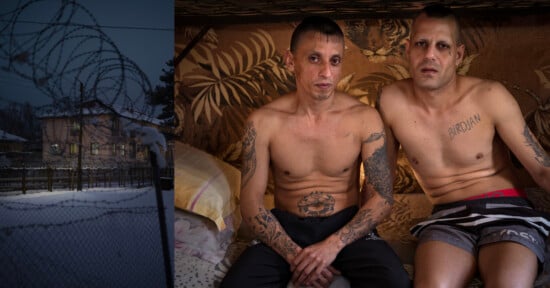 Two shirtless men with tattoos sit on a bed in a dimly lit room with animal print wallpaper; on the left, a snowy, fenced area with barbed wire and houses is visible.