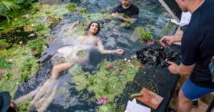 A woman in a white dress floats in a pond with lily pads and flowers, surrounded by film crew members and camera equipment during a photoshoot.