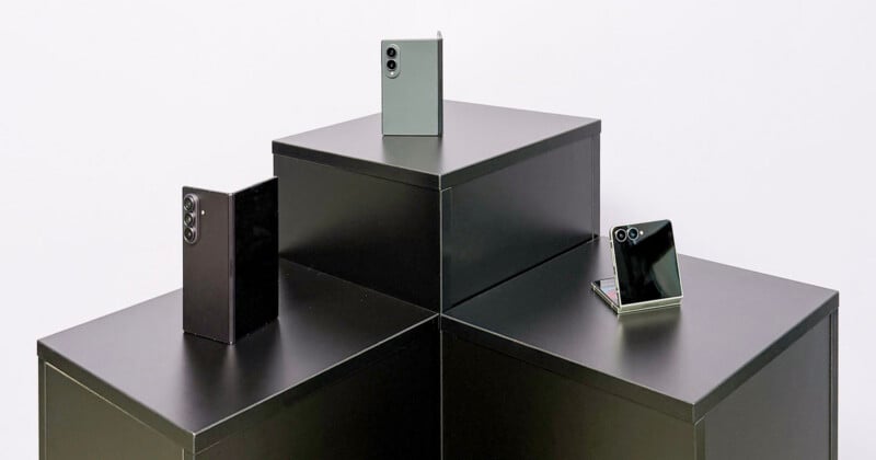 Three foldable smartphones are displayed on interconnected black podiums. Two phones are partially open, while one is fully open. All have dual rear cameras and reflective surfaces, set against a plain white background.