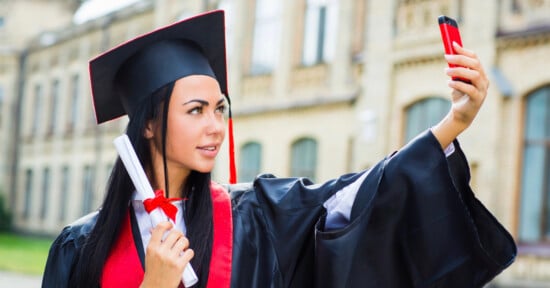A graduate in cap and gown holds a diploma and takes a selfie outside a building, smiling proudly.