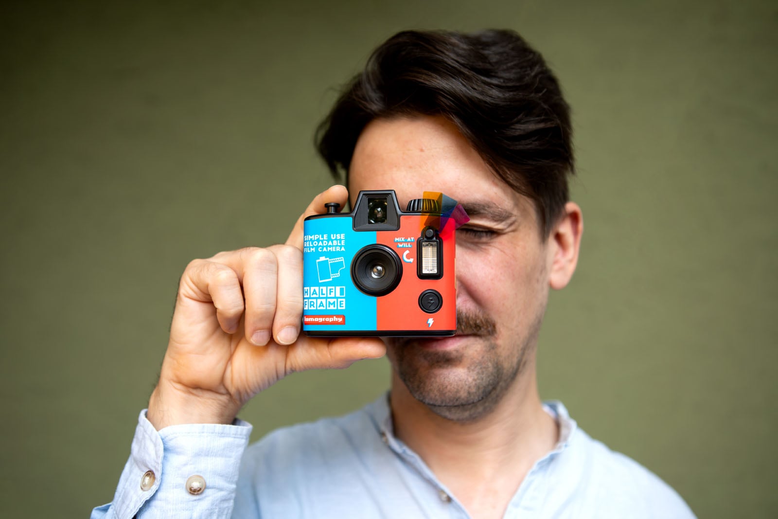 A person in a light blue shirt holds a colorful disposable camera up to their face and looks through the viewfinder, preparing to take a photo, against a plain background.