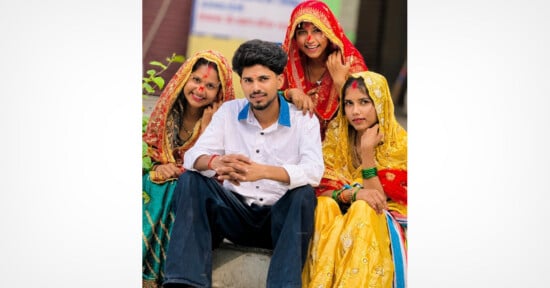 A young man sits on steps, surrounded by three smiling women in colorful traditional attire and veils, posing together outdoors.