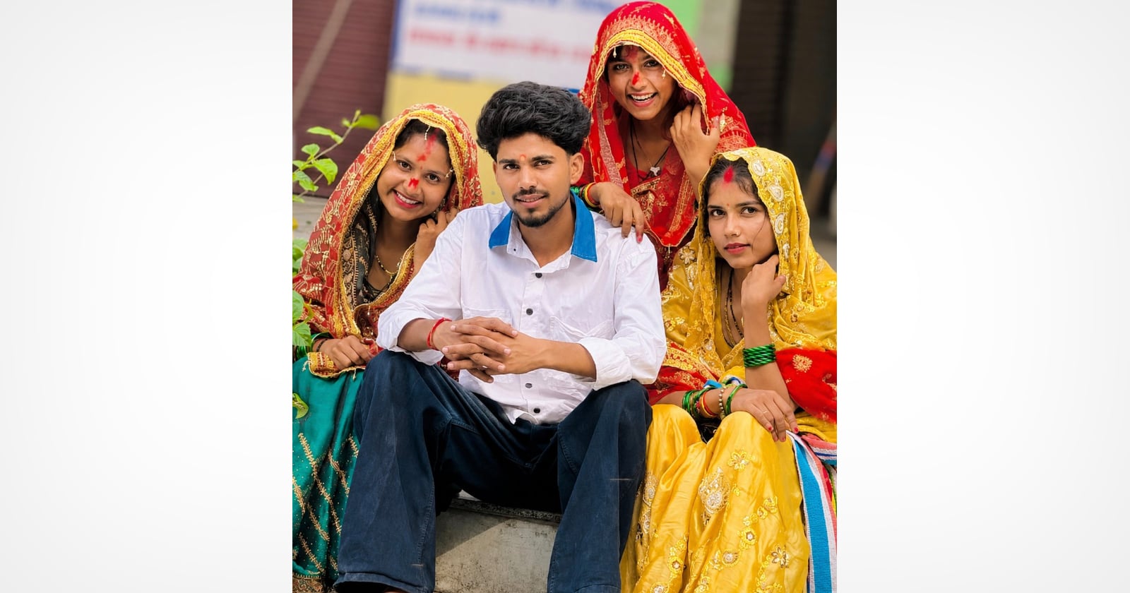 A young man sits on steps, surrounded by three smiling women in colorful traditional attire and veils, posing together outdoors.