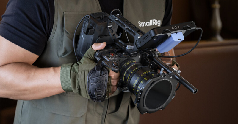 Person wearing a green vest and glove holds a professional video camera rig with various accessories attached, including a large lens and mounted external equipment.