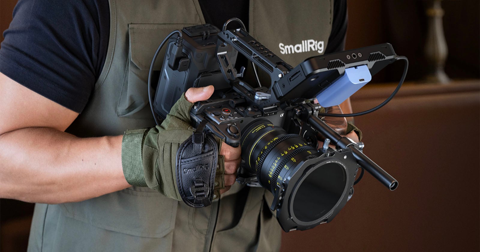 Person wearing a green vest and glove holds a professional video camera rig with various accessories attached, including a large lens and mounted external equipment.
