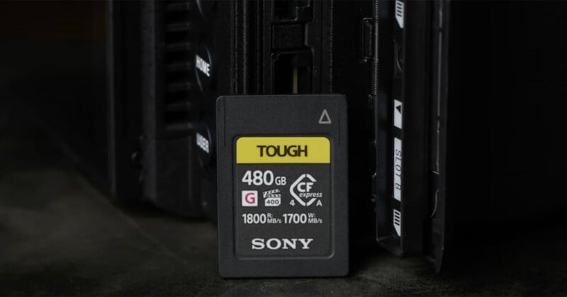 A Sony Tough CFexpress Type A memory card with 480GB capacity is standing upright in front of an electronic device, possibly a camera or card reader, on a dark surface.