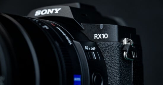 Close-up of a black Sony RX10 camera showing the textured grip, part of the zoom lens, focus mode switch, and the RX10 label against a dark background.