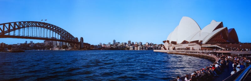 A wide-angle view of the Sydney Harbour Bridge and the Sydney Opera House across the water, with people sitting on a pier.