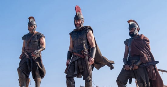 Three men dressed as ancient warriors in helmets and armor stand outdoors against a clear blue sky, holding swords and looking serious, portraying a historical or mythological scene.