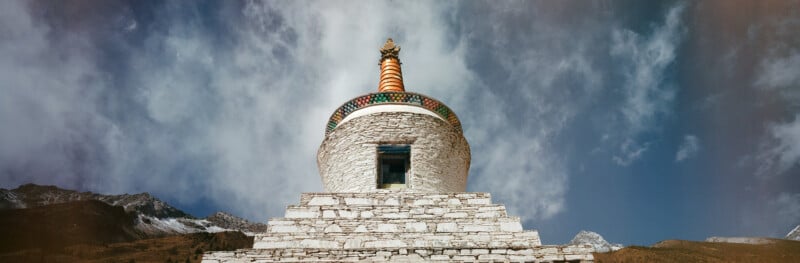 A white stone stupa with a golden spire sits against a cloudy blue sky, flanked by mountain peaks.