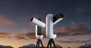 Two white, modern, robotic telescopes on tripods stand against a twilight sky with mountains in the background.