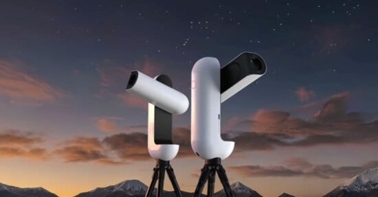 Two white, modern, robotic telescopes on tripods stand against a twilight sky with mountains in the background.