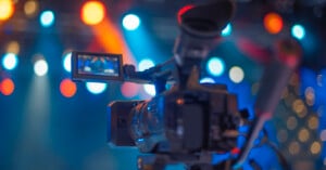 A close-up of a video camera filming in a colorful studio with bright, blurred stage lights in the background.
