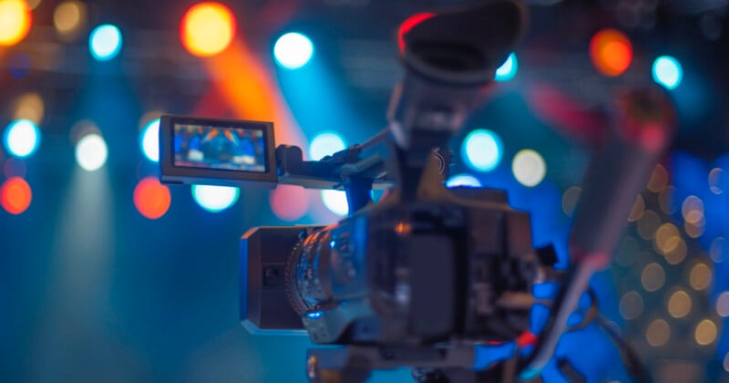 A close-up of a video camera filming in a colorful studio with bright, blurred stage lights in the background.