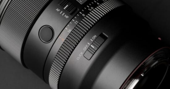 A close-up view of a camera lens barrel featuring an aperture ring with f-stop markings, an AF/MF switch, a customizable button, and a click on/off switch.