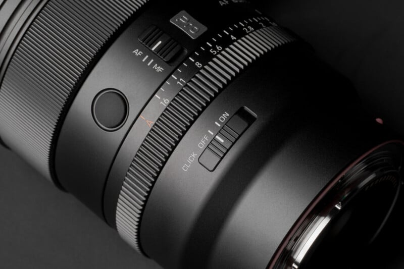 A close-up view of a camera lens barrel featuring an aperture ring with f-stop markings, an AF/MF switch, a customizable button, and a click on/off switch.