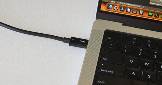 A Thunderbolt cable is plugged into a port on a laptop, next to the keyboard and screen showing application icons. The setup is on a white surface.