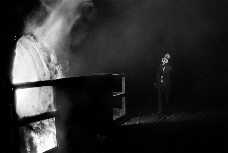 A person wearing protective gear and goggles stands near a large, bright furnace or molten material in a dark industrial setting.