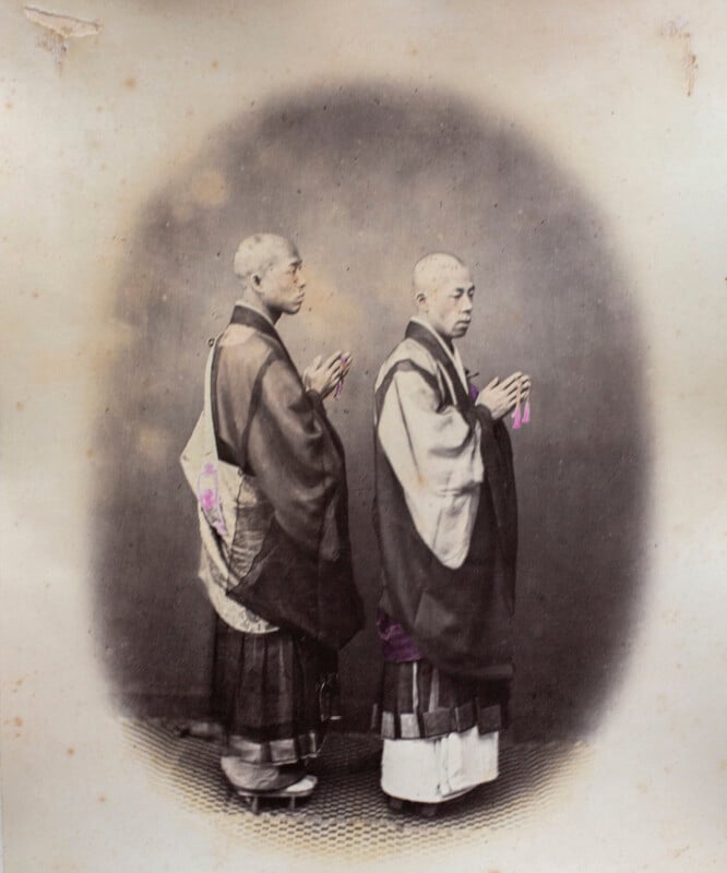 Two Japanese monks with shaved heads stand in profile, wearing traditional robes and holding prayer beads in their hands.
