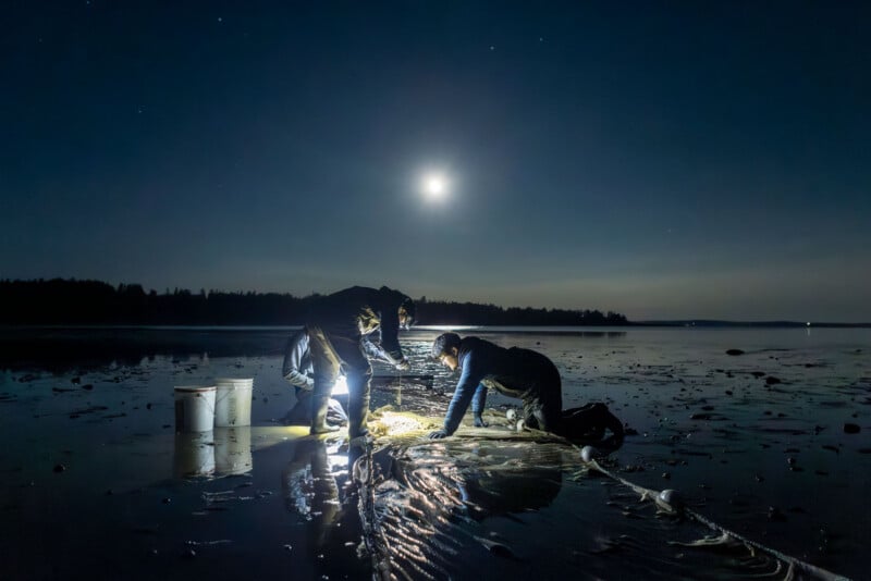 Two people in dark clothing kneel on a wet, muddy shore at night, illuminated by a bright moon and a small light while tending to a fishing net.