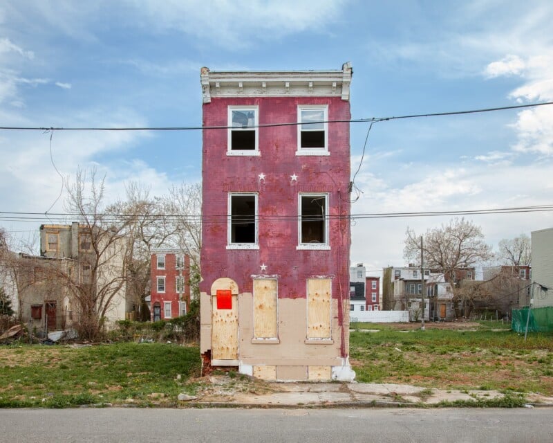 A narrow, three-story brick row house stands isolated in an empty lot, with boarded-up windows and a red no-trespassing sign.