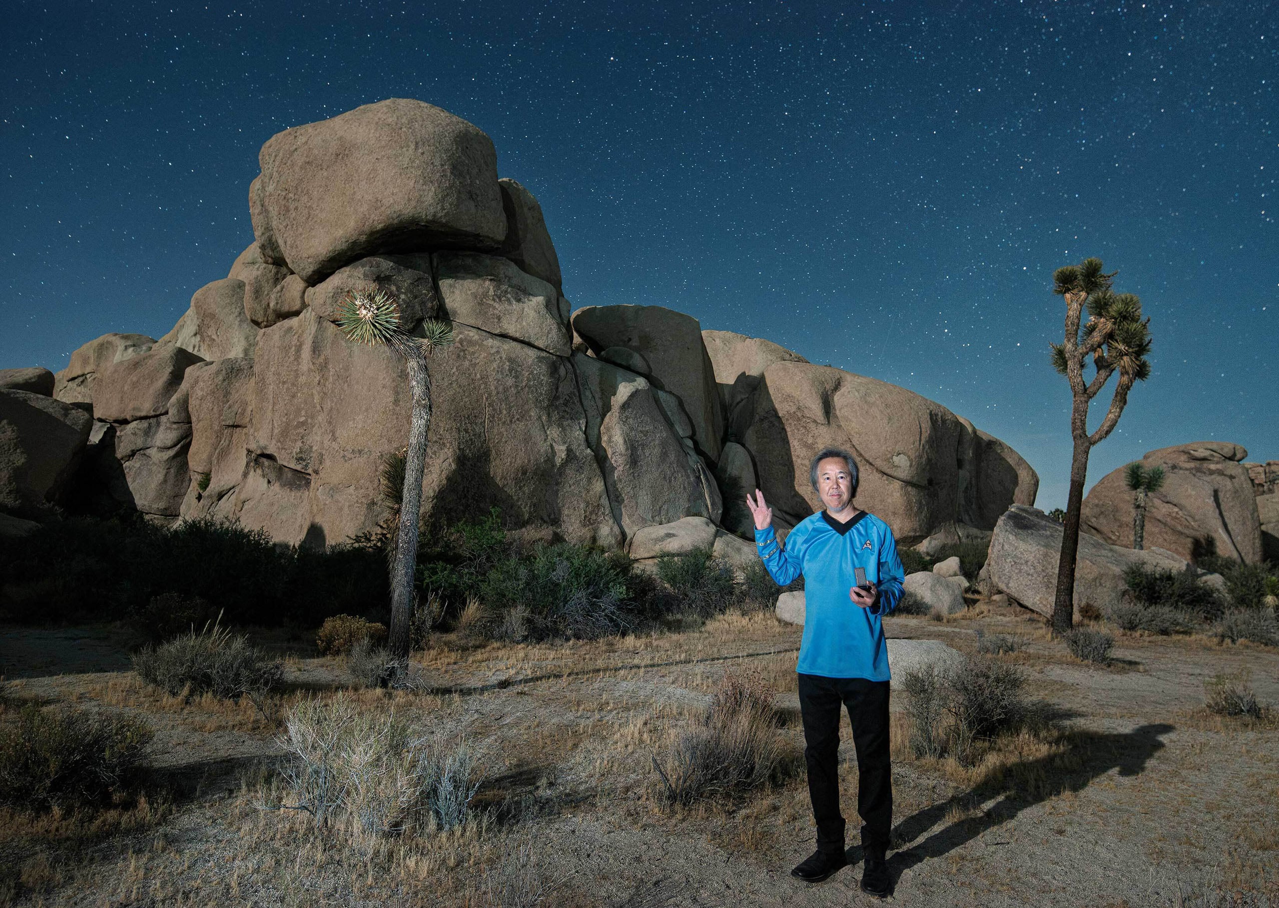 A man wearing a blue Star Trek-style uniform stands in a desert landscape at night, making a Vulcan salute gesture under a starry sky.